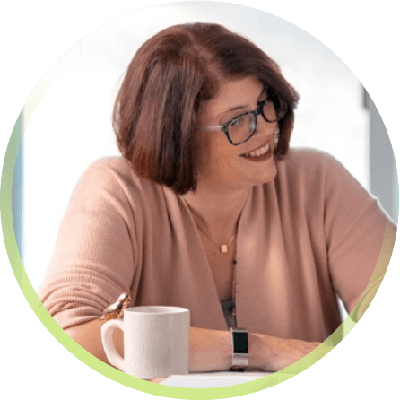 A white woman with short brown hair and patterned glasses is seen from the chest up. She looks off camera to the right, smiling. In front of her is a white coffee mug