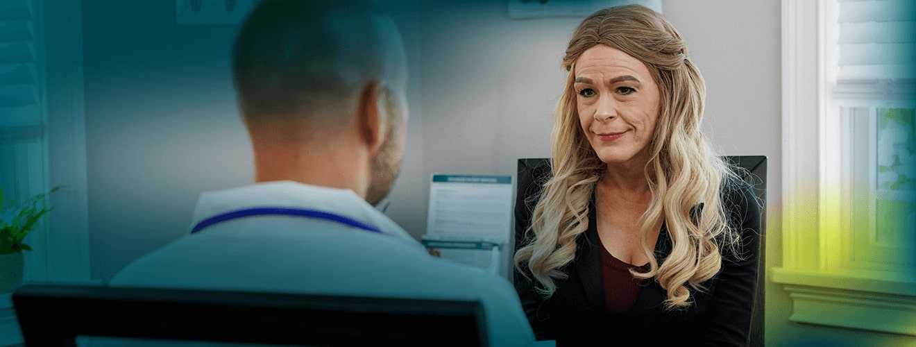 A white woman with long blonde hair is seen from the chest up. She is seated and talking to a male doctor who is seen from behind.
